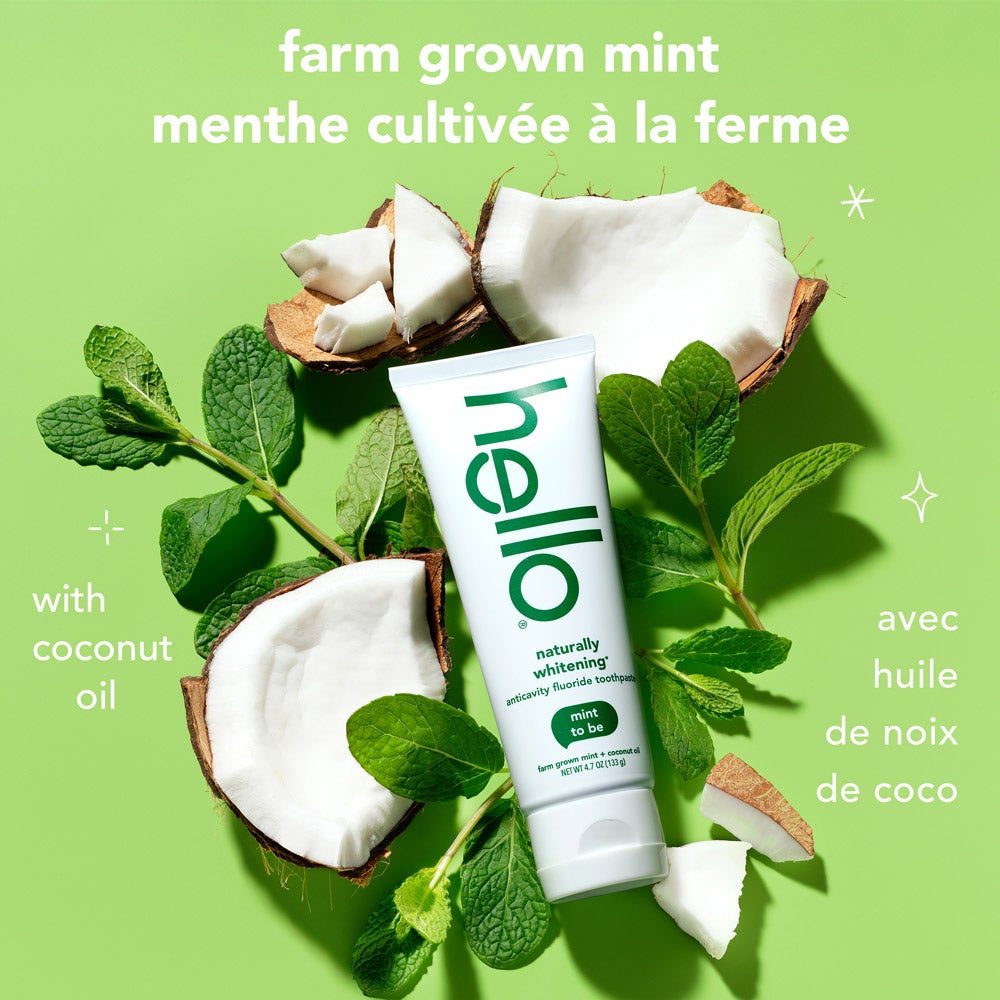 A tube of hello naturally whitening fluoride toothpaste with farm grown mint stands upright among fresh mint leaves and cracked coconut pieces on a light green background. Text above the tube says, "farm grown mint" in English and French, and "with coconut oil" in both languages.